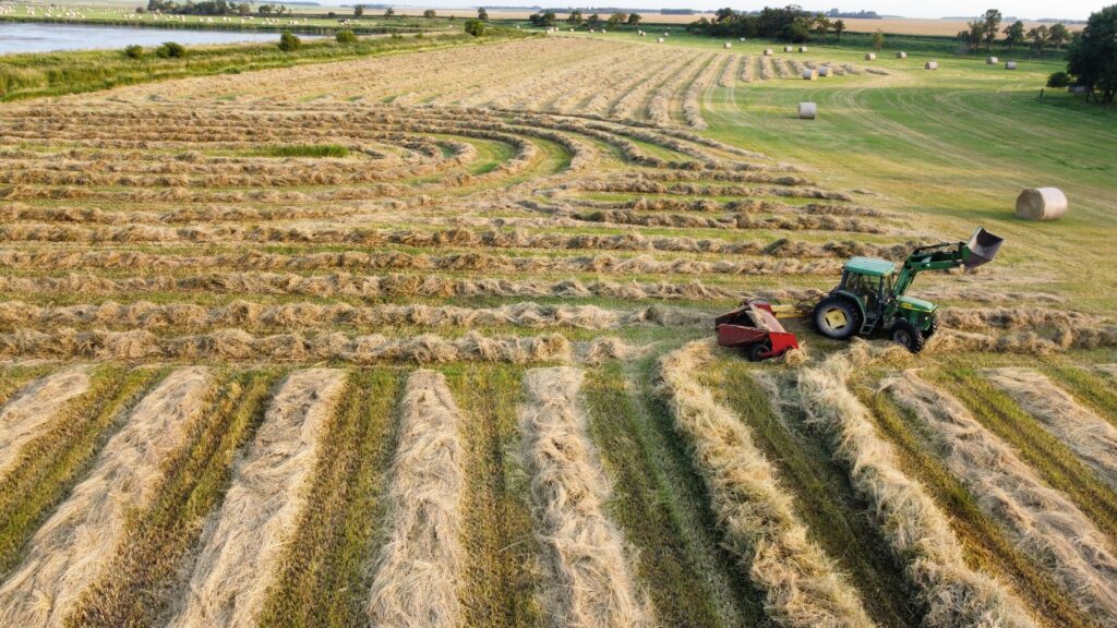 Aerial view of a hay field with curved windrows and a green tractor pulling equipment, surrounded by scattered round hay bales and open farmland.