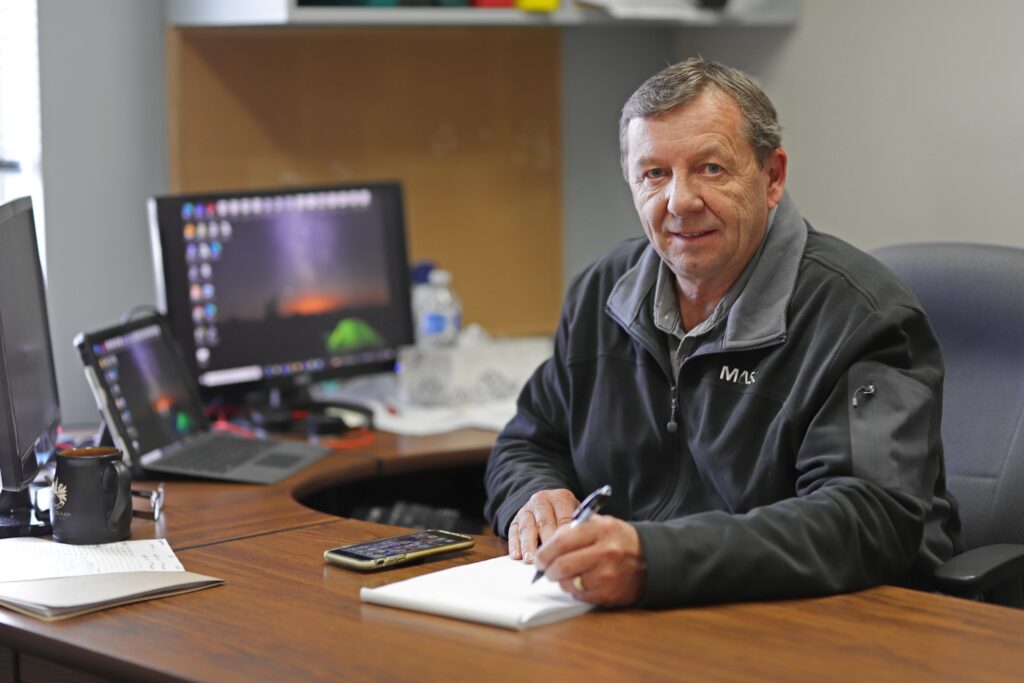 MASC staff seated at an office desk writing in a notebook, with a computer monitor, laptop, calculator, and coffee mug on the desk.