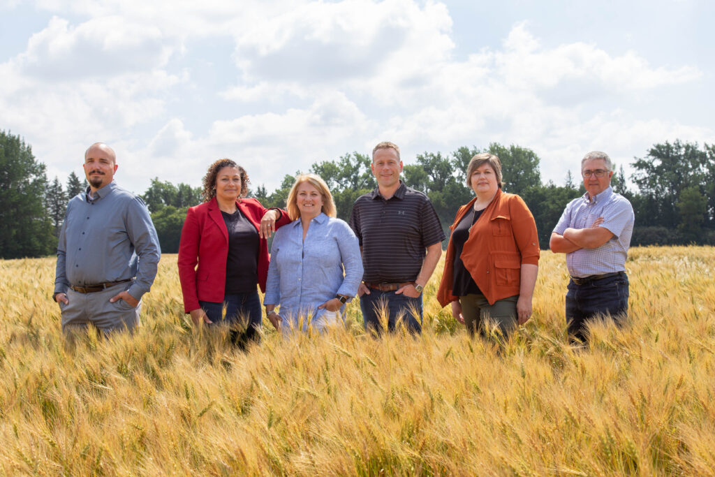 Members of MASC’s Executive Management Committee standing together in a golden wheat field, wearing casual and business attire, with trees and a bright sky in the background.