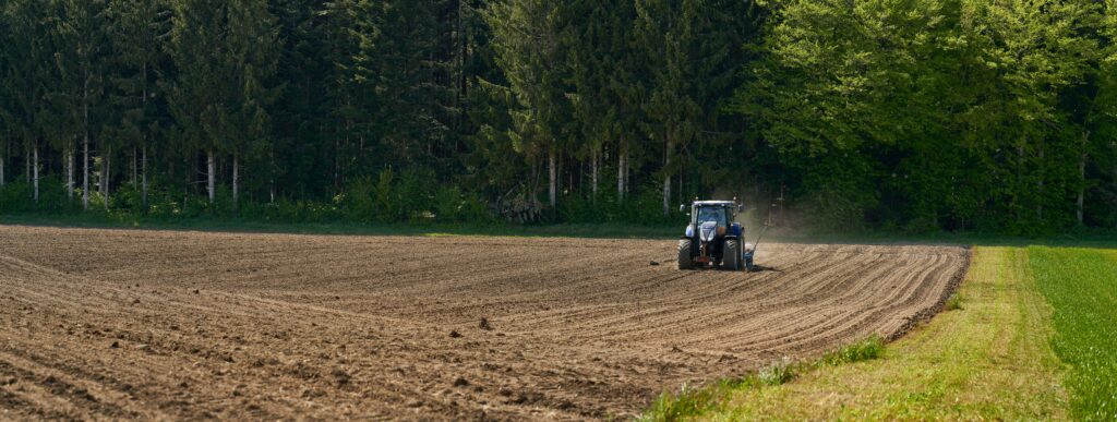 Vue plongeante d’un tracteur vert dans un grand champ brun en labour, traçant des sillons parallèles sur le sol.