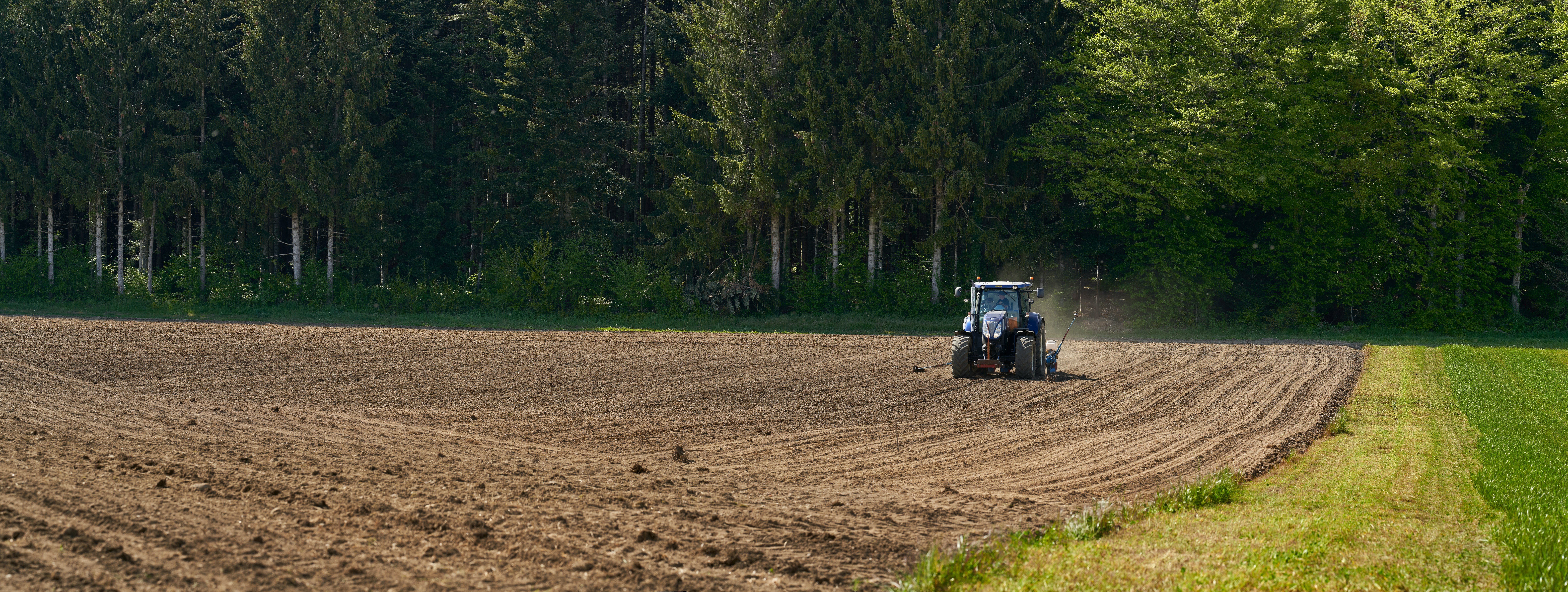 Vue plongeante d’un tracteur vert dans un grand champ brun en labour, traçant des sillons parallèles sur le sol.